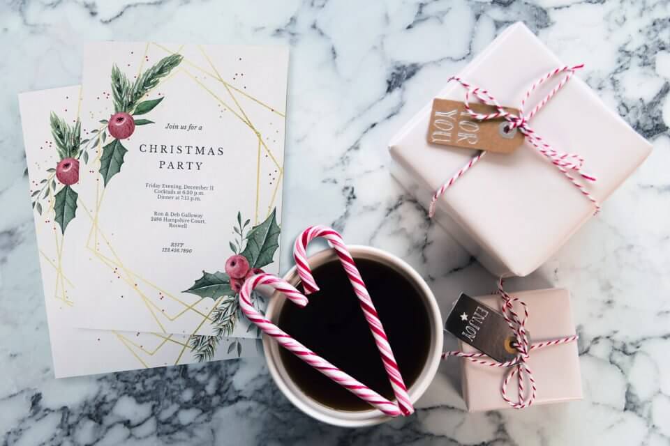 Top view of a Christmas party table with invitations, hot cocoa, and gifts. The invitation reads 'Join us for a Christmas party' with a white background, adorned with Christmas greenery illustration and a golden frame.