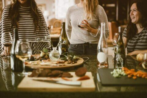 Three women enjoying a Friendsgiving party, chatting and savoring drinks. The table is beautifully set with a spread of food and beverages. Cover for the blog post 'How to Plan the Perfect Friendsgiving Party'.