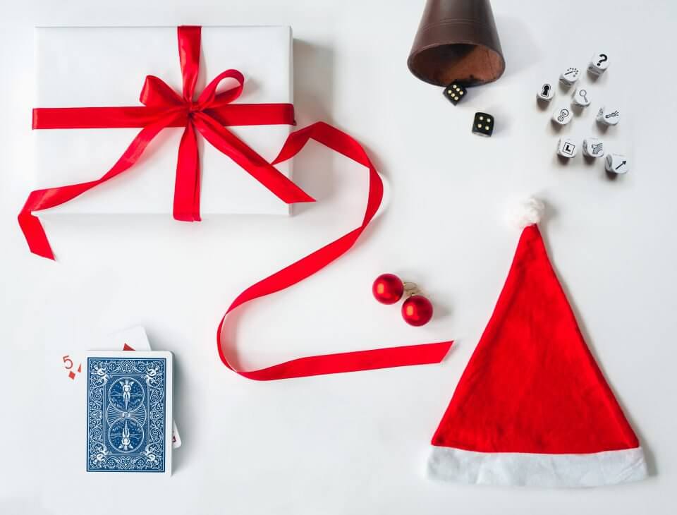 Flat lay view of Christmas party objects on a table: a white-wrapped gift with a red bow, game dice with figures, a Santa hat, Christmas ornaments, and playing cards.