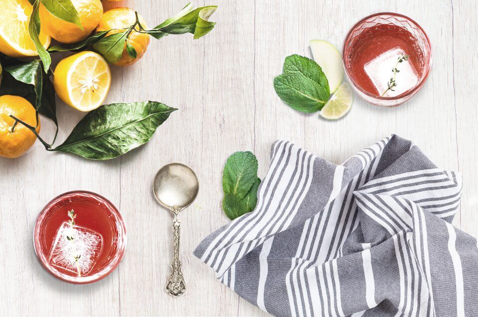 Galentine's Day Cocktails: Two Glasses with Red Drinks, Ice Cubes, Measuring Spoon, Lemons, and Kitchen Towel on a Wooden Surface.