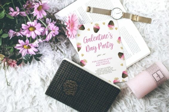 Galentine's Day invite with chocolate-dipped strawberries illustration, set on a white plush surface amid an open book, watch, flowers, notebook, and unlit candle. Perfect for Your Ultimate Guide to Hosting a Memorable Galentine’s Day Party.