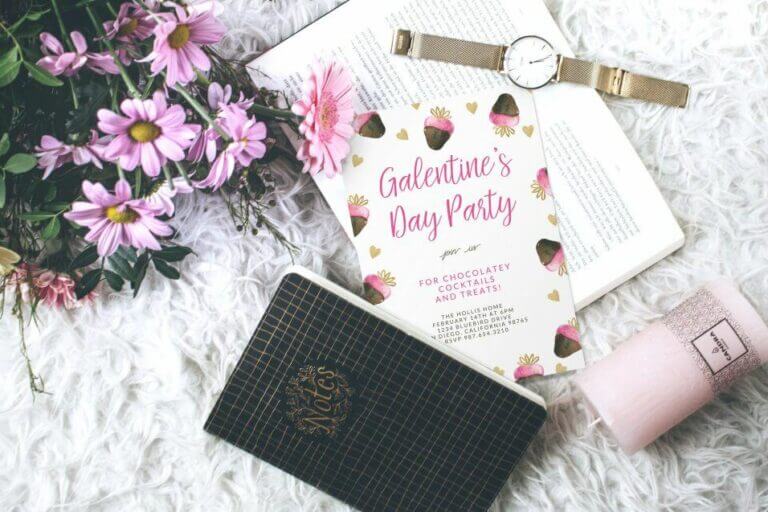 Galentine's Day invite with chocolate-dipped strawberries illustration, set on a white plush surface amid an open book, watch, flowers, notebook, and unlit candle. Perfect for Your Ultimate Guide to Hosting a Memorable Galentine’s Day Party.