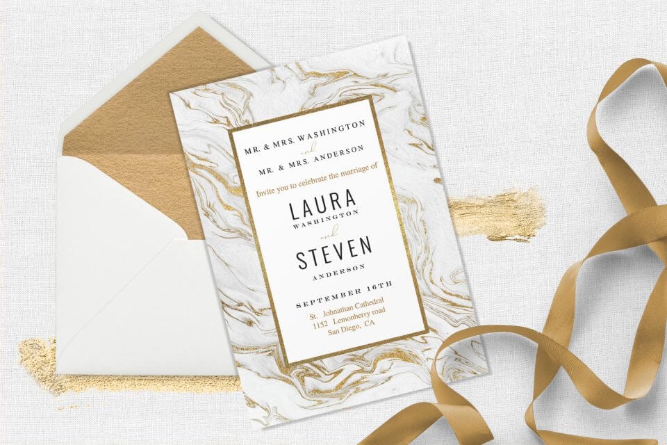 Marble-themed wedding invitation with gold accents, set on a white and gold-lined envelope, adorned by a nearby golden ribbon.