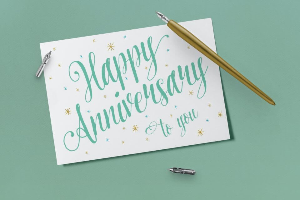 Elegant script in teal and gold conveying warm wishes for a Happy Anniversary. The sentiment 'Happy Anniversary to You' gracefully placed on a green surface alongside a pen, adding a touch of sophistication to the celebration