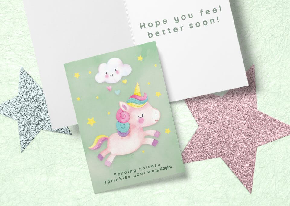 A 'Best Get Well Wishes & Quotes For A Speedy Recovery' greeting card, featuring a whimsical unicorn illustration, is placed on a flat surface. Alongside the card are two sparkly star cut-outs, adding a magical touch to the setup. An open note lies next to the card