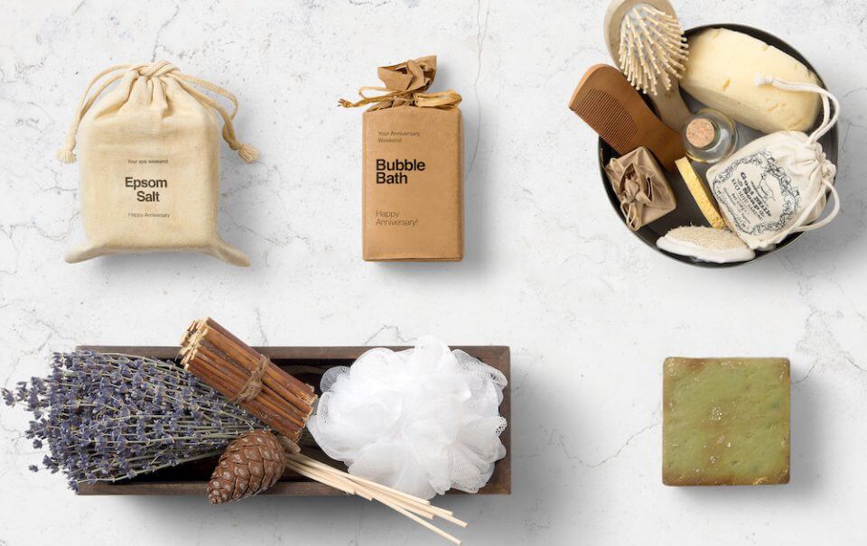 A serene spa arrangement designed to captivate with romance, featuring a bag of Epsom salt, a bag of bubble bath, and a tray adorned with lavender, cinnamon sticks, and a showering sponge. Adjacent, a basket holds hair care essentials