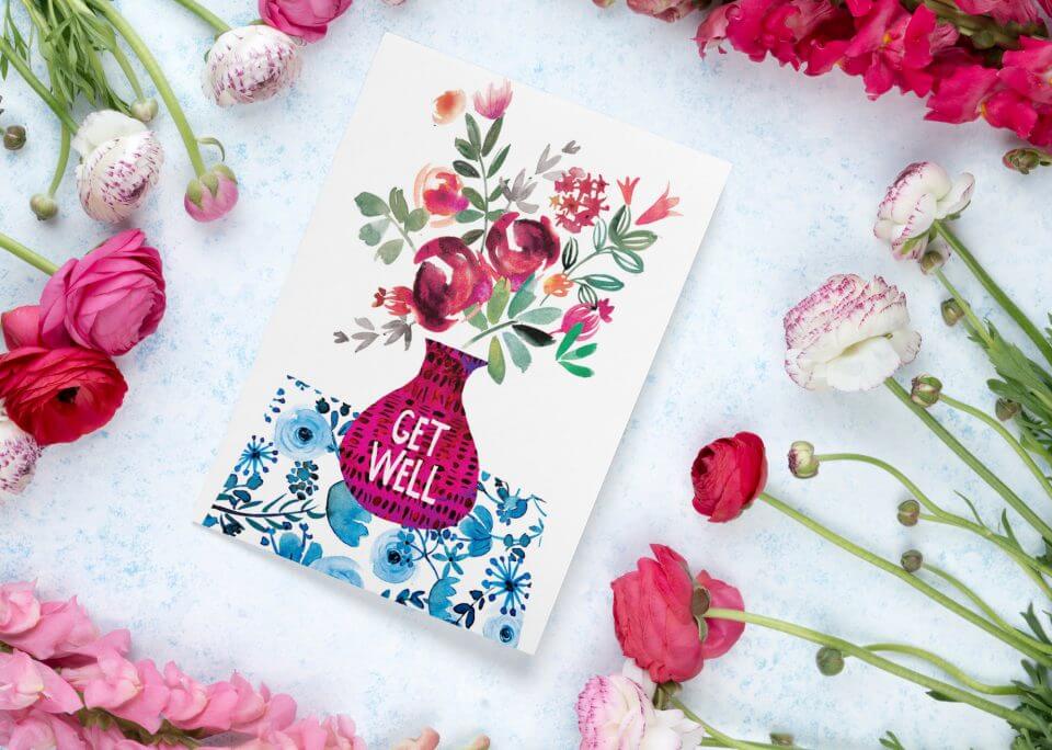 A 'Best Get Well Wishes & Quotes For A Speedy Recovery' greeting card, adorned with an illustration of a pink vase filled with vibrant flowers, is elegantly placed on a surface. Surrounding the card is an abundant arrangement of assorted flowers, creating a lush and colorful display