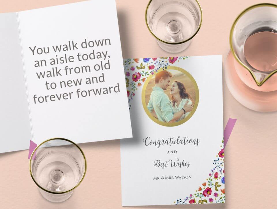 Charming cameo-style wedding card featuring floral illustrations, a gold ring with a couple's photo, and the words 'Congratulations' and 'Best Wishes.' Rests on a pink surface alongside an open card and champagne glasses.