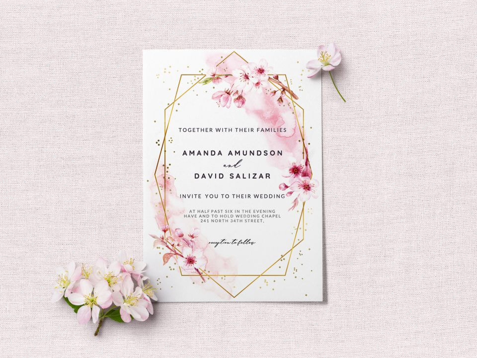 Cherry blossom wedding invite: Elegant gold geometric border, adorned with blossoms. Black text. Rests on a light pink surface, surrounded by real cherry blossoms.