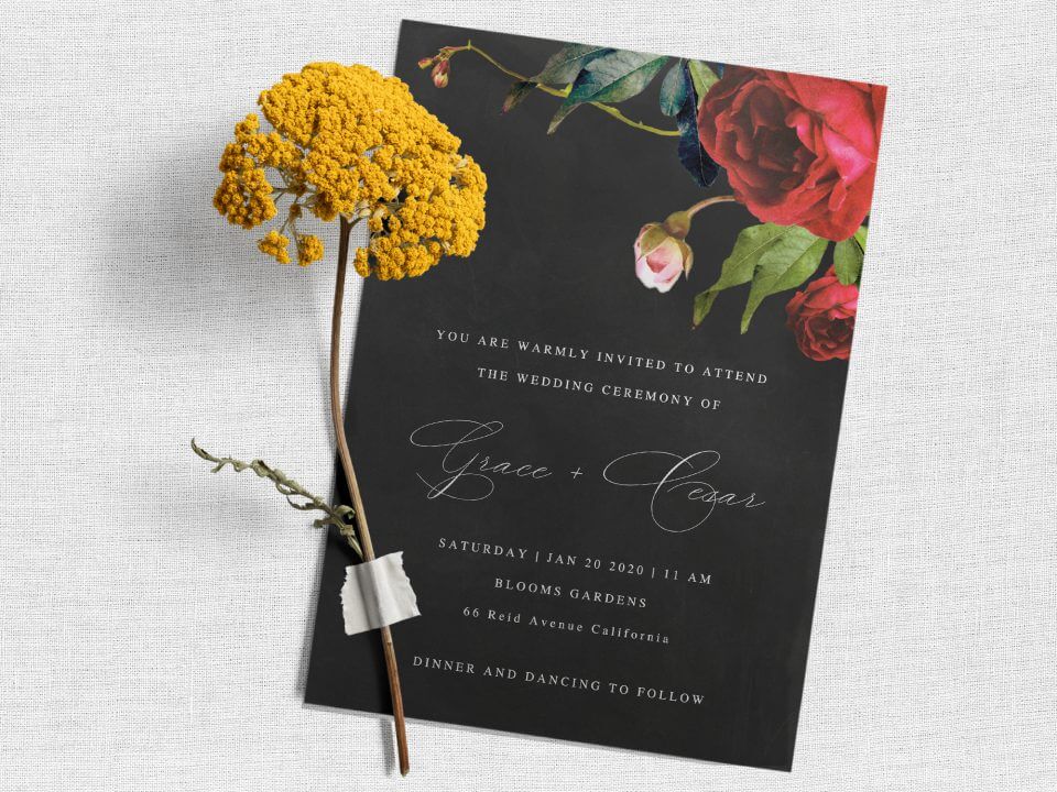 Climbing Roses Wedding Invite: Black background with rose illustration, white text. Rests on light grey surface with a vibrant yellow flower atop