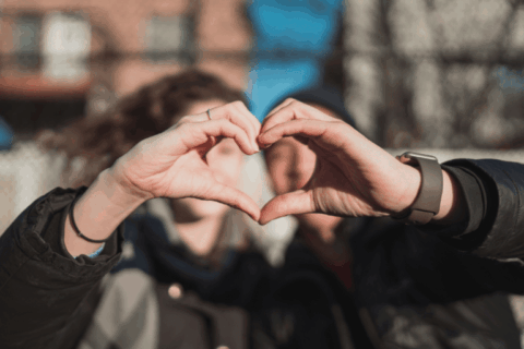 Two people forming a heart shape with their hands in the foreground, with a softly blurred couple standing together outdoors in the background.