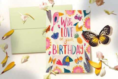 Vibrant Birthday Card: 'With Love on Your Birthday' in Multicolored Text, Adorned with Illustrations of Flowers, Butterflies, and Birds. Resting on a Neutral Background with a Light Green Envelope, Flower Petals, and a Striking Black & Yellow Butterfly. Perfect Cover for the Blog Post: '60+ Heartfelt Ways to Wish 'Happy Birthday' to Your Beloved One'