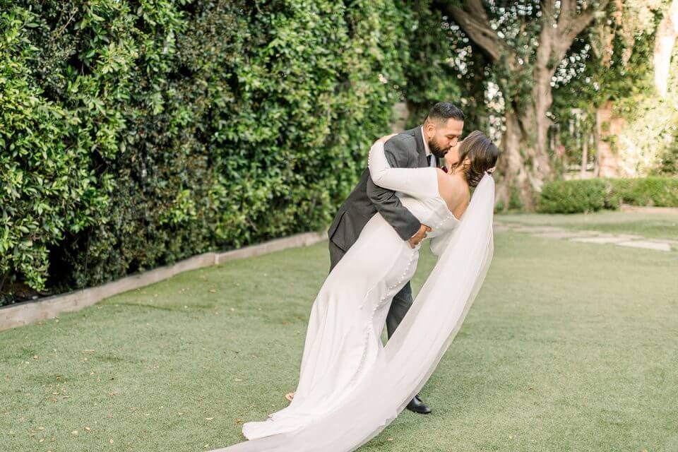 Enchanting Garden Vow Renewal: Groom tenderly embraces his bride in a white dress, sharing a romantic kiss amidst a sunlit garden