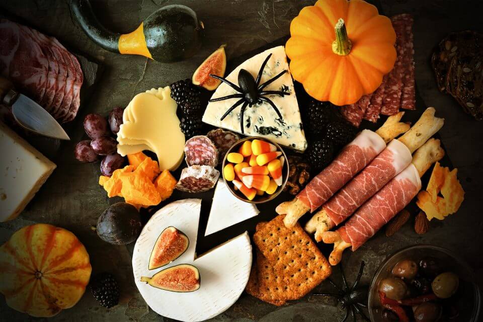 Halloween Charcuterie Board with Brie, Cheddar, Blue, and Goat Cheeses, Assorted Crackers, Fruits, and Vegetables. Festively Decorated with a Small Pumpkin and Plastic Spiders.