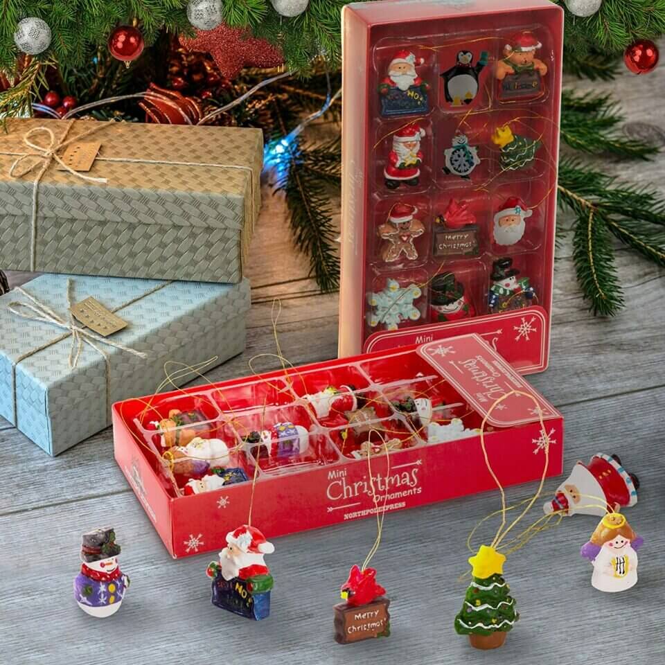 Assorted Christmas tree ornaments including snowflakes, snowmen, Santas, and penguins neatly arranged in a box. The boxes are placed at the base of a festively decorated Christmas tree, accompanied by two beautifully wrapped gifts