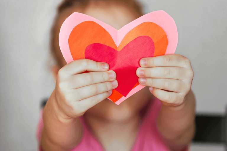 Capturing the spirit of love with a sweet image of a girl holding up a heart-shaped paper crafted from three different-sized hearts. Cover for '15 Valentine’s Day Party Ideas for Kids That Will Spread the Love'