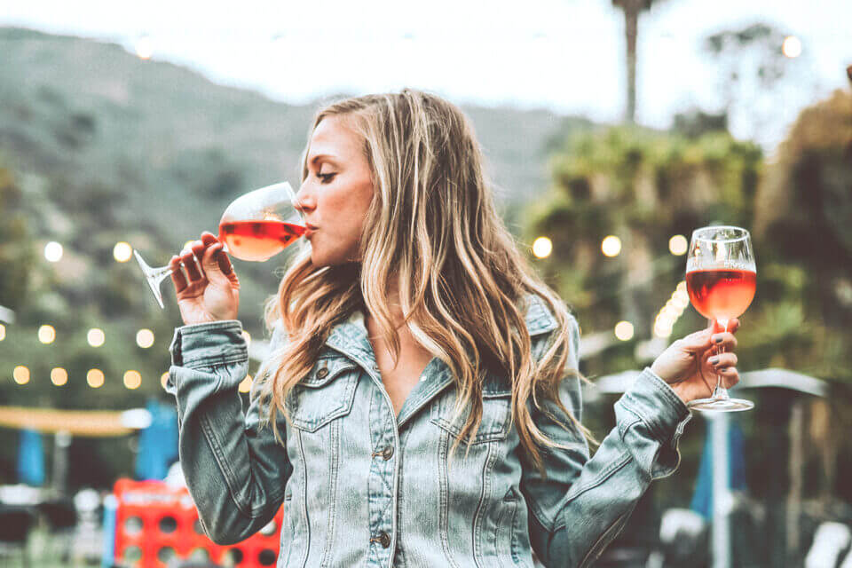 Blonde Girl Enjoying Wine Outdoors, Holding Two Glasses, Taking a Sip