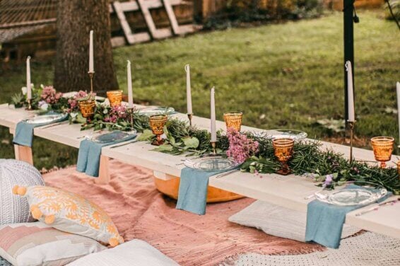 Enchanting Spring Outdoor Gathering: A Beautifully Decorated Low Table Adorned with a Long Centerpiece of Lush Flowers and Greenery, Illuminated by Elegant Candlesticks and Complemented by Vibrant Orange Glasses. Comfortable Seating Arrangements with Plush Pillows