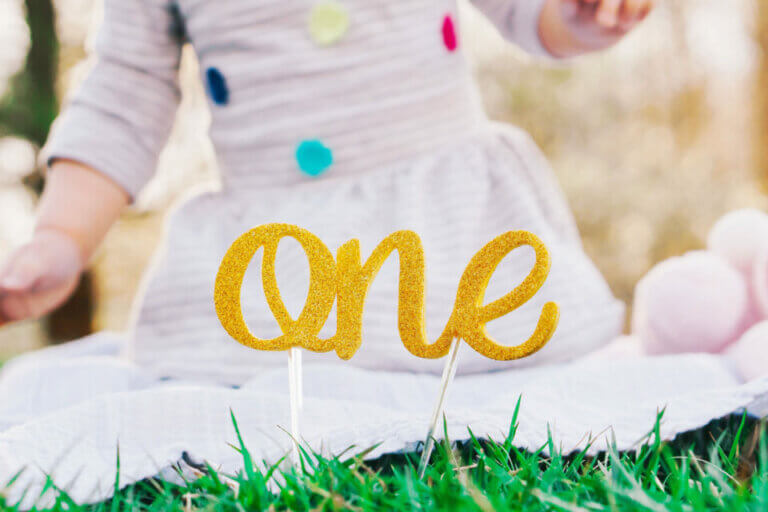 Golden 'One' Sign Graced by a Toddler in a Cheerful Polka-Dotted White Dress. A Charming Cover for '20 of the Most Popular First Birthday Party Themes'.