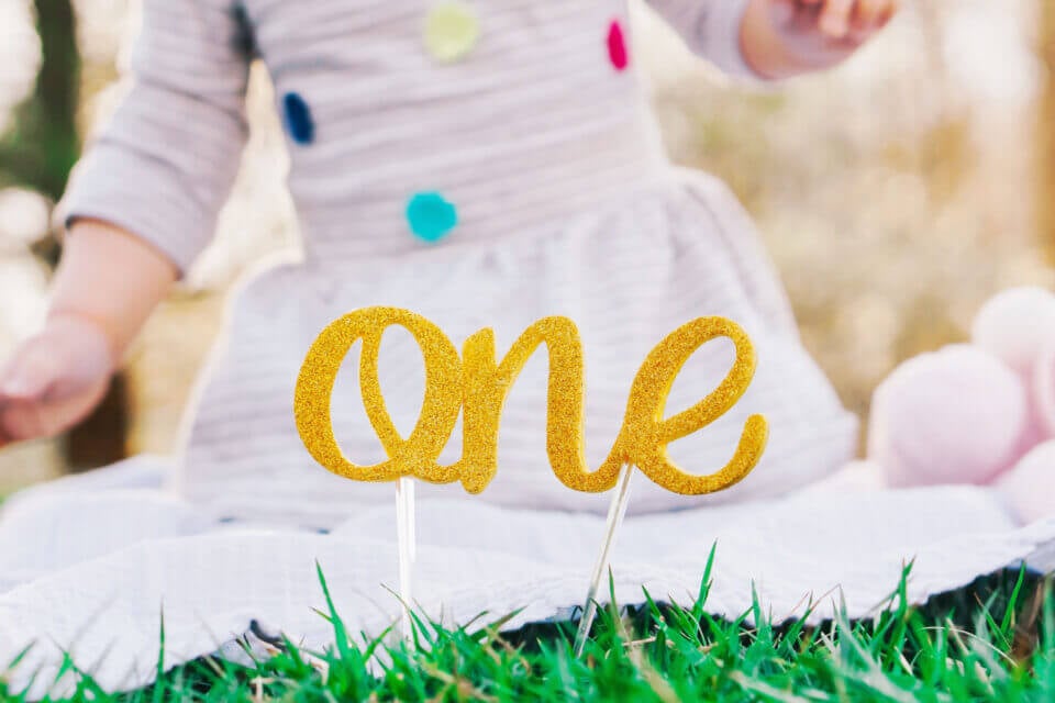 Golden 'One' Sign Graced by a Toddler in a Cheerful Polka-Dotted White Dress. A Charming Cover for '20 of the Most Popular First Birthday Party Themes'.