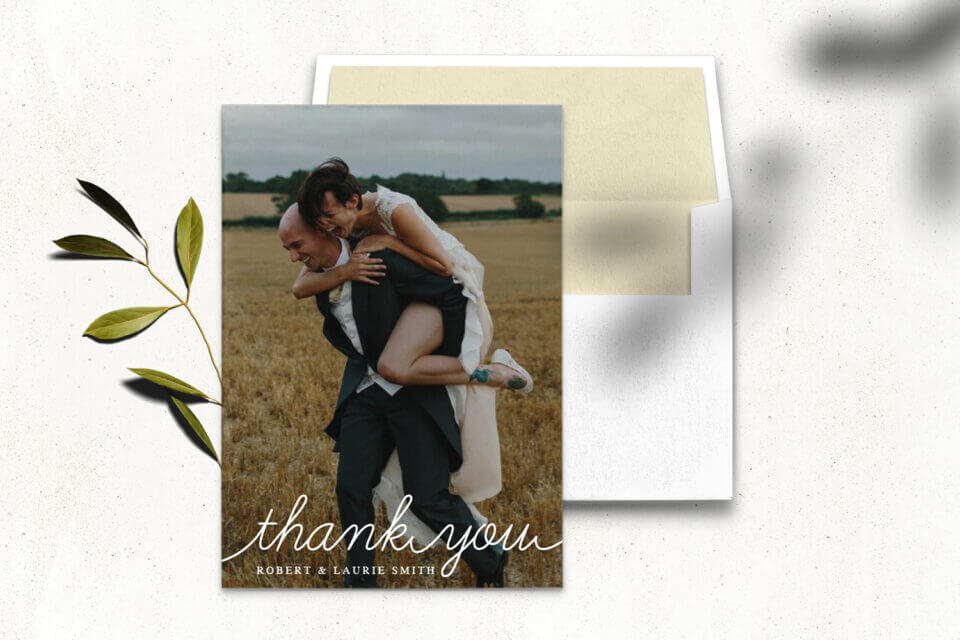 Bride joyfully hops on groom's back, both laughing. 'Thank You' in white over the image on the wedding card. Rests on an open white envelope, adorned with a nearby flowering branch.