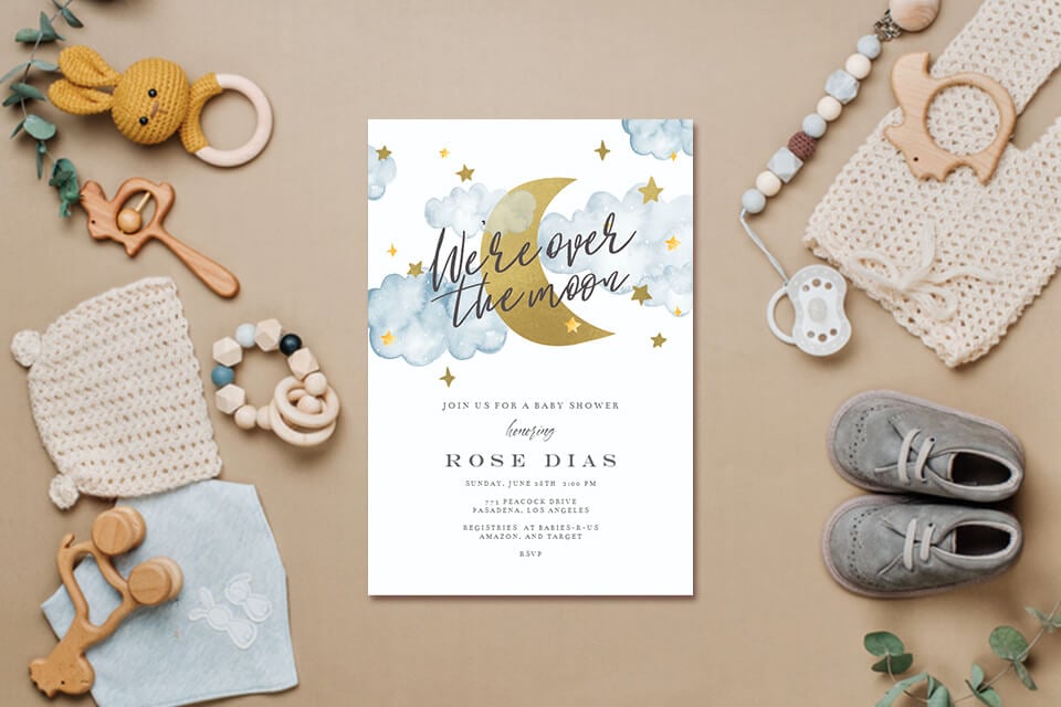 Gender-Neutral Baby Shower Themes: 'Over the Moon' Invitation with Celestial Delights. Featuring a golden moon amidst light blue clouds and shimmering stars. Accompanied by a delightful flat lay of baby essentials including shoes, toy, rattle, and knitted attire.