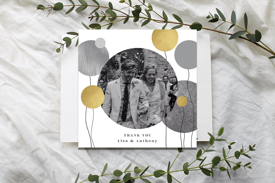 Wedding thank you card with balloon illustrations and a central couples' photo. Resting on a white sheet, flanked by two tree branches for a natural touch.