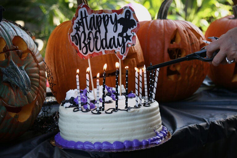 Enchanting Halloween Birthday Cake: White with Purple Accents, Aglow with Lit Candles, and a Festive 'Happy Booday' Topper. Background Features Artfully Carved Pumpkins. Cover for '21 Spectacular Halloween Birthday Party Ideas to Bewitch Your Guests'