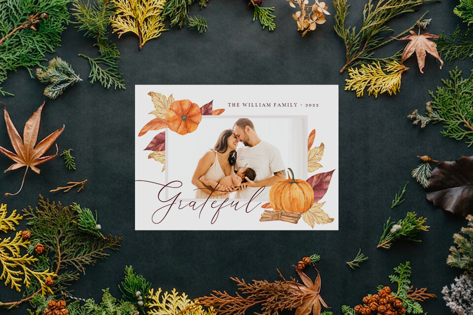 Thanksgiving card featuring a cherished family photo and the word 'Grateful'. The invite is adorned with beautifully illustrated golden autumn leaves, resting on a tabletop embellished with pine tree branches, evoking warmth and gratitude.