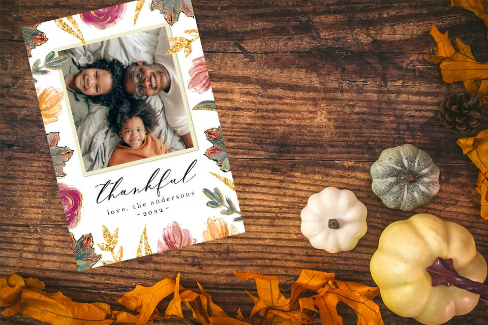 Thanksgiving card featuring a family of three with the word 'Thankful' inscribed. The card is elegantly displayed on a rustic wooden surface adorned with a trio of pumpkins in varying sizes, complemented by vibrant autumn leaves