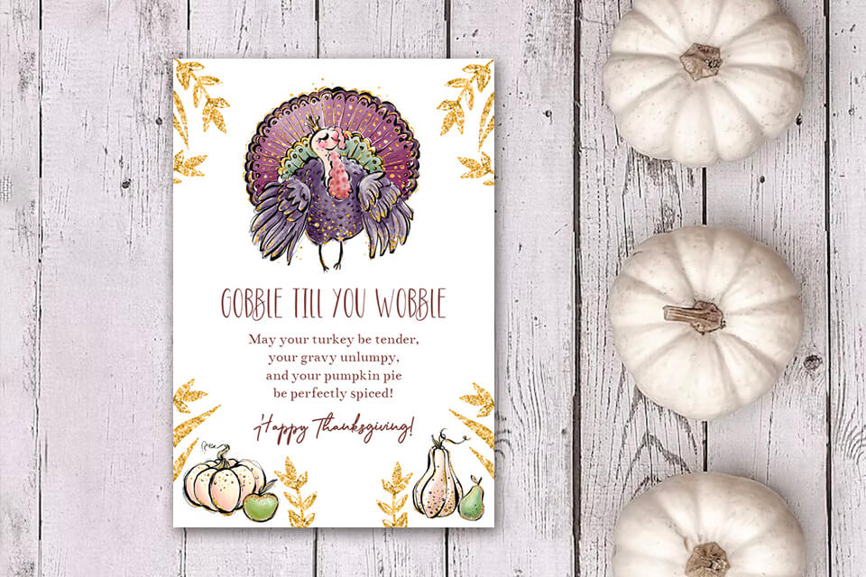 Thanksgiving card with a whimsical 'Gobble Till You Wobble' message and a charming peacock illustration. The card is displayed on a rustic wooden surface adorned with three white pumpkins, adding a touch of elegance to the festive atmosphere.