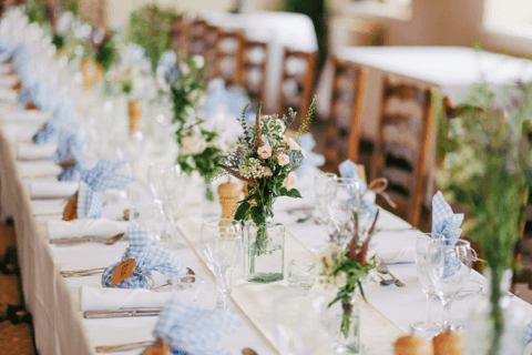 A beautifully arranged wedding reception table set with elegance and sophistication. The white tablecloth complements the wooden chairs, adorned with vases of fresh flowers. Natural light floods the room through large windows, creating a warm and inviting atmosphere for a memorable wedding rehearsal dinner. Blog post: '10 Wedding Rehearsal Dinner Ideas Your Friends and Family Will Actually Love'