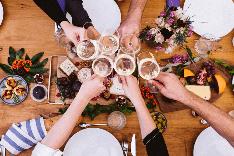 Overhead view capturing a moment of celebration, with people's hands raising wine glasses in a cheer over a table laden with food, epitomizing the joy of a party gathering.