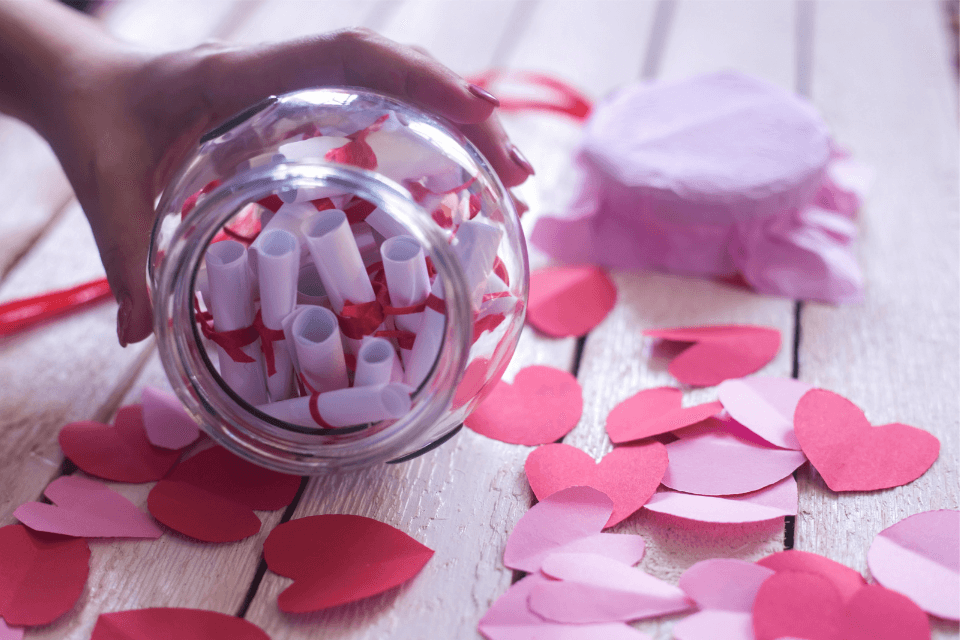 person holding a glass jar filled with little scrolls of paper placed on a table with little paper hearts scattered around