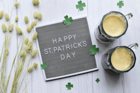 Cheerful St. Patrick's Day message written on a rustic chalkboard, set upon a wooden surface beside glasses of beer, accented with three delicate clover leaves for festive decor.