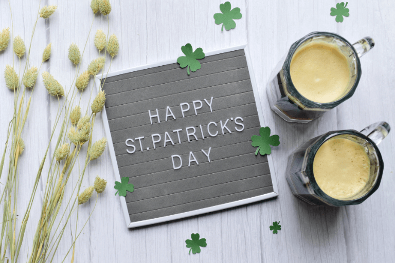 Cheerful St. Patrick's Day message written on a rustic chalkboard, set upon a wooden surface beside glasses of beer, accented with three delicate clover leaves for festive decor.