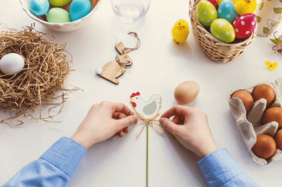 Hands delicately arranging Easter decorations: a chicken centerpiece being adjusted while nearby, a wooden rabbit decor and baskets filled with colorful eggs complete the festive scene
