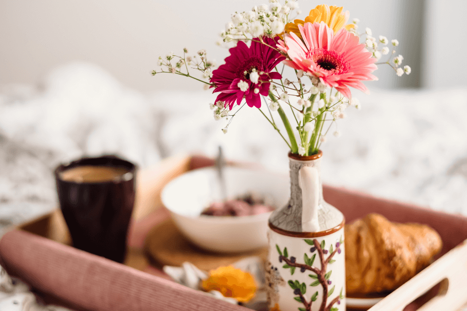 A close-up photograph of breakfast in bed, with the focus on a vase of fresh flowers in the foreground, adding a touch of elegance to the morning.