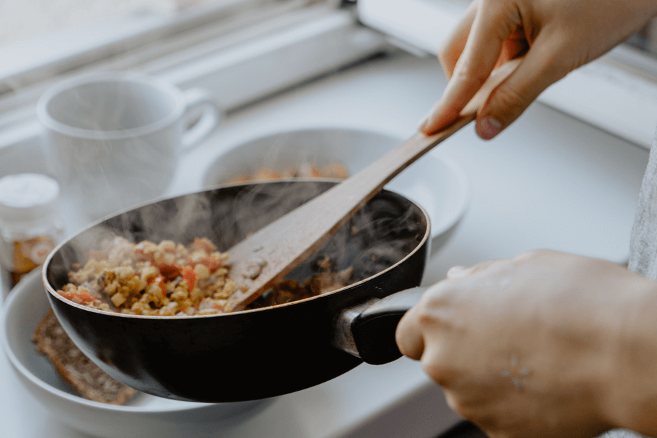 A woman prepares a home-cooked meal, stirring ingredients in a cooking pan with a wooden spoon, the kitchen filled with the aroma of comfort food.