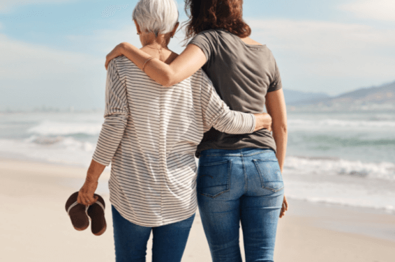 From behind, an older mother and her adult daughter walk along the beach, hands tenderly wrapped around one another, enjoying the day's gentle embrace.