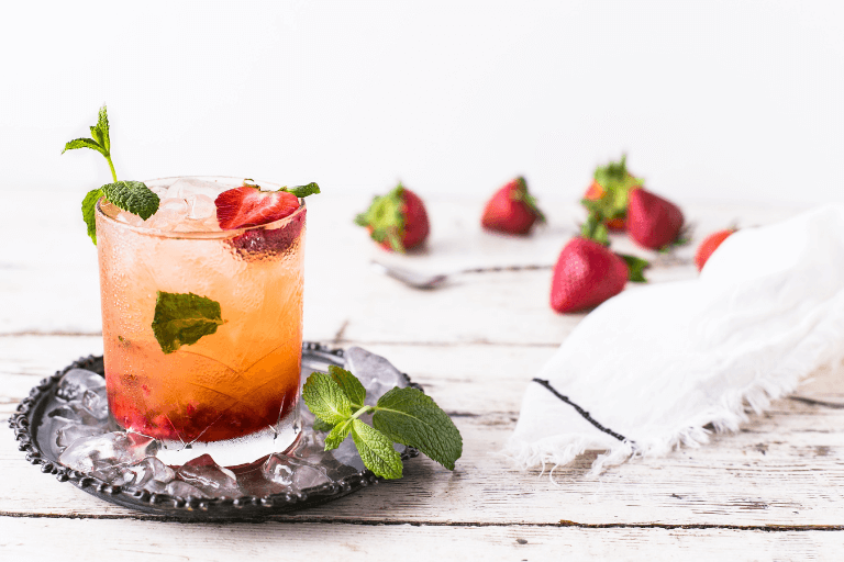 A vibrant cocktail with orange slices, fresh mint leaves, and strawberries in a glass, set on a wooden surface with scattered strawberries nearby, inviting a taste of summer freshness.