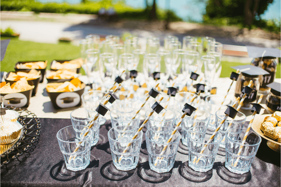 A close-up photograph of a graduation party table décor, featuring drink glasses elegantly decorated with straws topped with miniature graduation hats. 