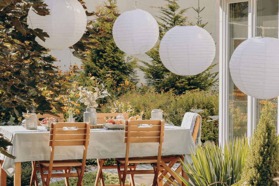 An outdoor home party venue bathed in sunlight, festively decorated with colorful paper lanterns, a beautifully set table, and wooden chairs inviting guests to celebrate on this cheerful day.