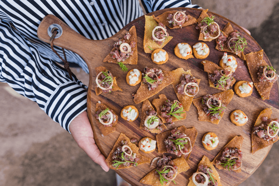 Overhead view of a delectable finger food platter at a party, with a person's hands gracefully carrying the wooden tray amongst the gathering.