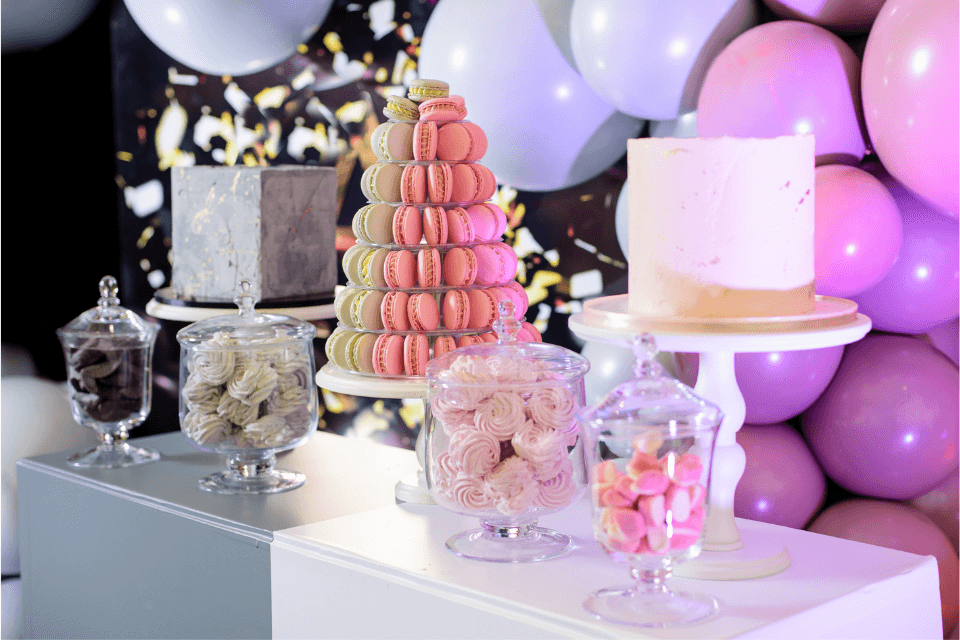 Charming birthday table setting in pastel hues, featuring an assortment of sweets. Macarons stand tall on a cake stand, flanked by two uniquely designed cakes. Glass containers brim with delectable treats. A festive balloon backdrop completes the delightful scene.