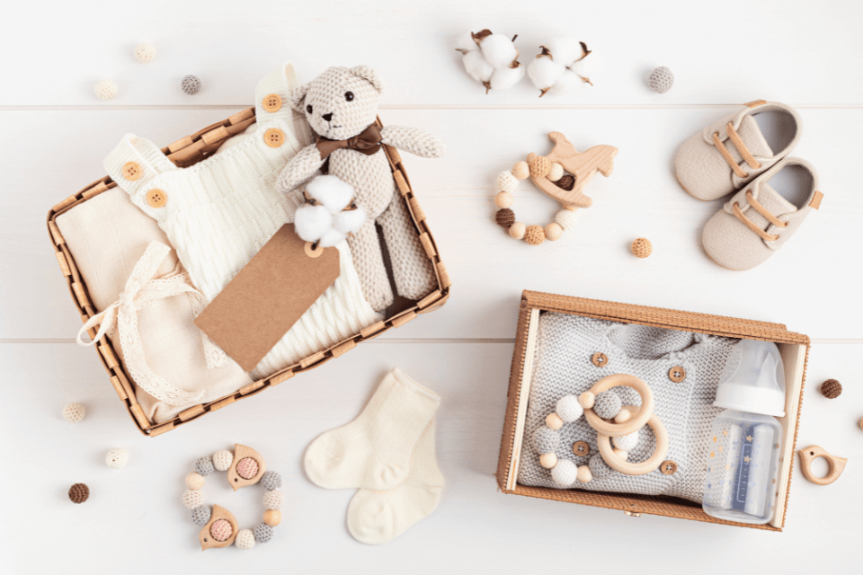 From above, a gender reveal party's baby gift baskets are showcased. One basket holds a small teddy bear and a onesie, while the other contains a bottle, jumper, and rattle. Nearby, two rattles, a pair of baby shoes, and socks complete the charming display.