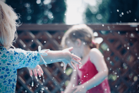 Blurred image of two young girls gleefully playing with water during an outdoor summer birthday party. Cover for the blog post 'Fun in the Sun: Exciting Themes for Kids' Summer Birthdays.