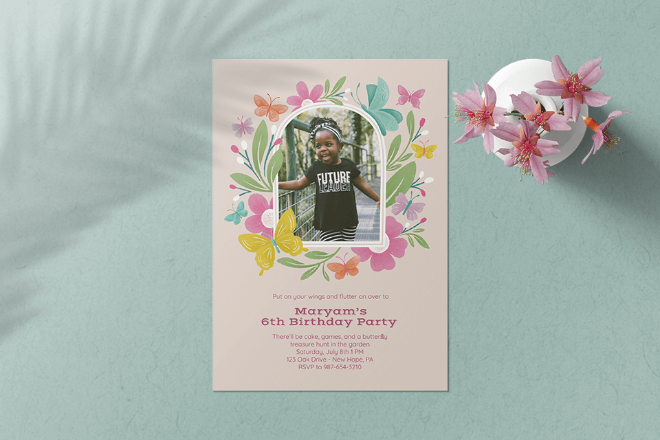 Birthday invite by Gia Graham with a child's photo, floral and butterfly border, on a green surface with pink flowers.