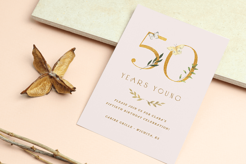 50th birthday party invitation to a '50 Years Young' celebration featuring an elegant illustration of the number 50 adorned with delicate butterflies. The invite is gracefully displayed on a table alongside a charming anise flower.