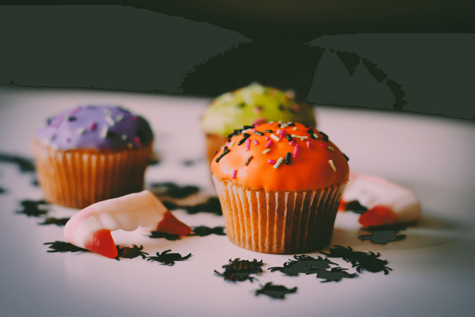 Three cupcakes, decorated with green, orange, and purple frosting, sit atop a table. Vampire gummy fangs and small spider cutouts surround the cupcakes, creating a spooky Halloween display.
